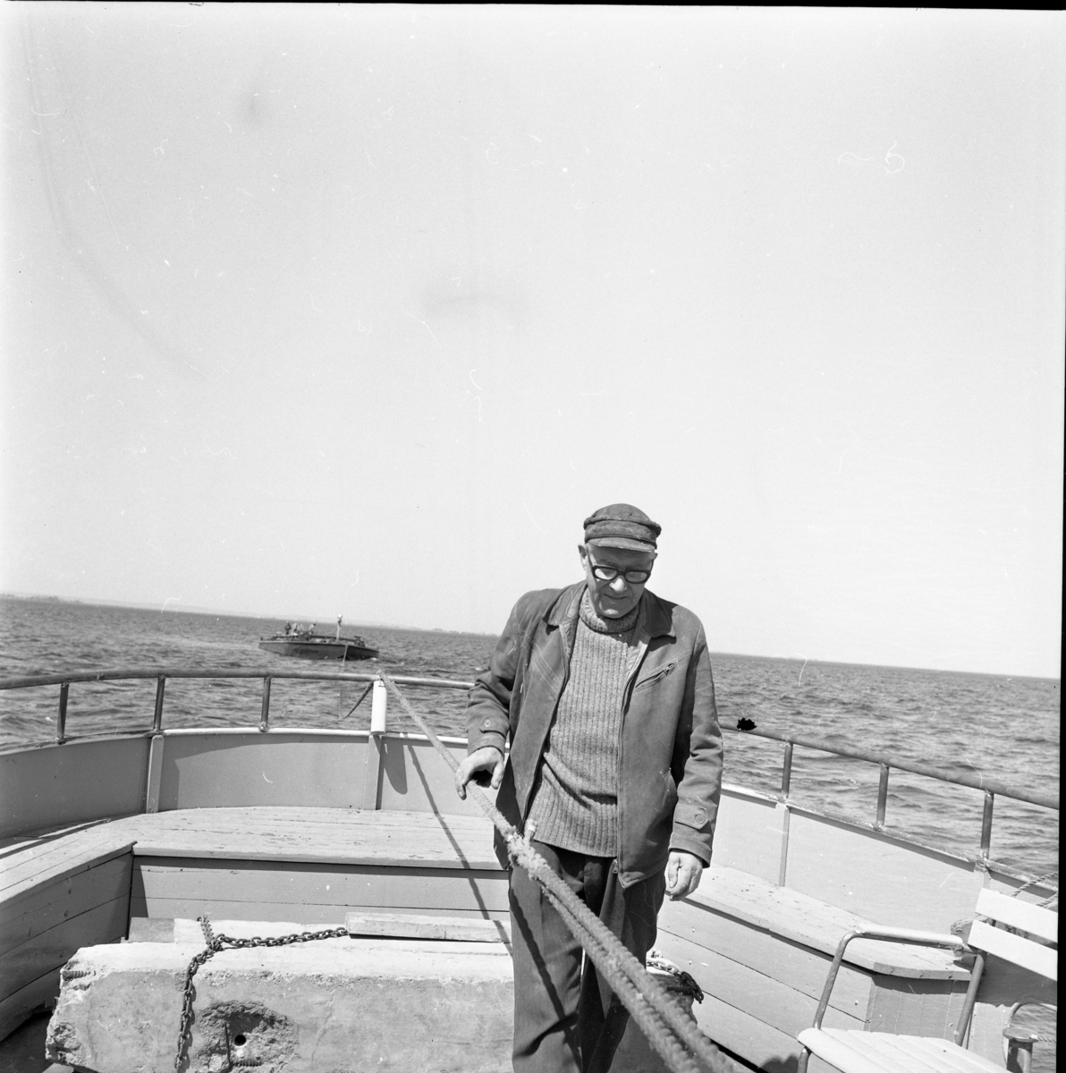 En man i glasögon och arbetskläder står i aktern till en båt, troligen Hebe II. På släp har man en pråm.