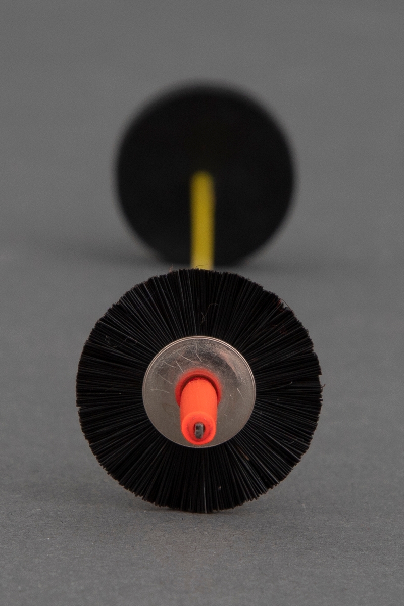 Brosje laget av en gul strikkepinne. På den ene enden er det påtredd et rødt plastrør. Mellom plasten er det påsatt et rundt metallhjul med svarte børster. På den andre enden, der spissen er, er det festet et rundt, svart gummihjul som kan taes av og settes på. Dette er lukkemekanismen til brosjen.