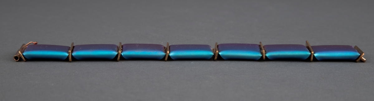 Leddet armbånd av titan. Leddene består av kvadratiske plater i magenta og blågrønt med en skimrende overflate. På baksiden er hjørnene brettet inn mot hverandre slik at leddene blir "pute"-formet. Kvadratene er tredd inn på to tynne metallkjettinger og med rektangulære gullfargede plater mellom hvert ledd. Låsen består av en metallpinne som stikkes ned i et rør.