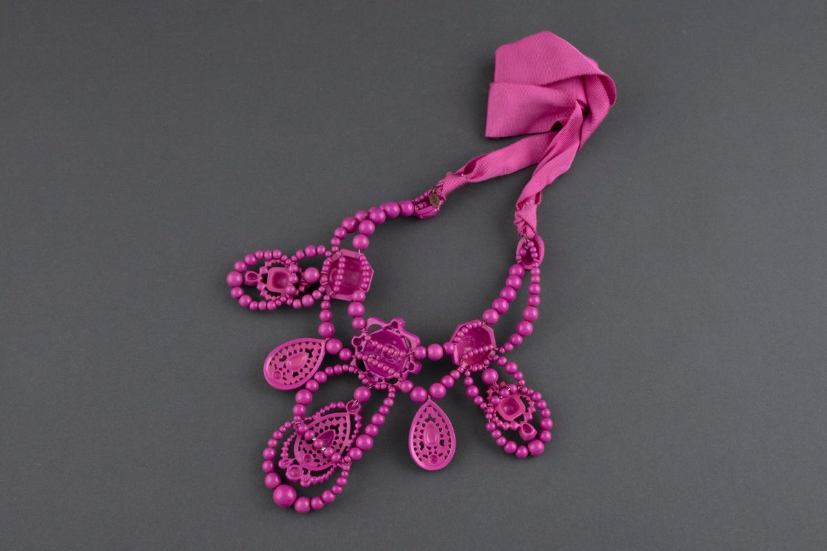 Stort halssmykke av rosalakkert metall, plastperler og brede tekstilbånd i samme farge. Smykket knytes i nakken.