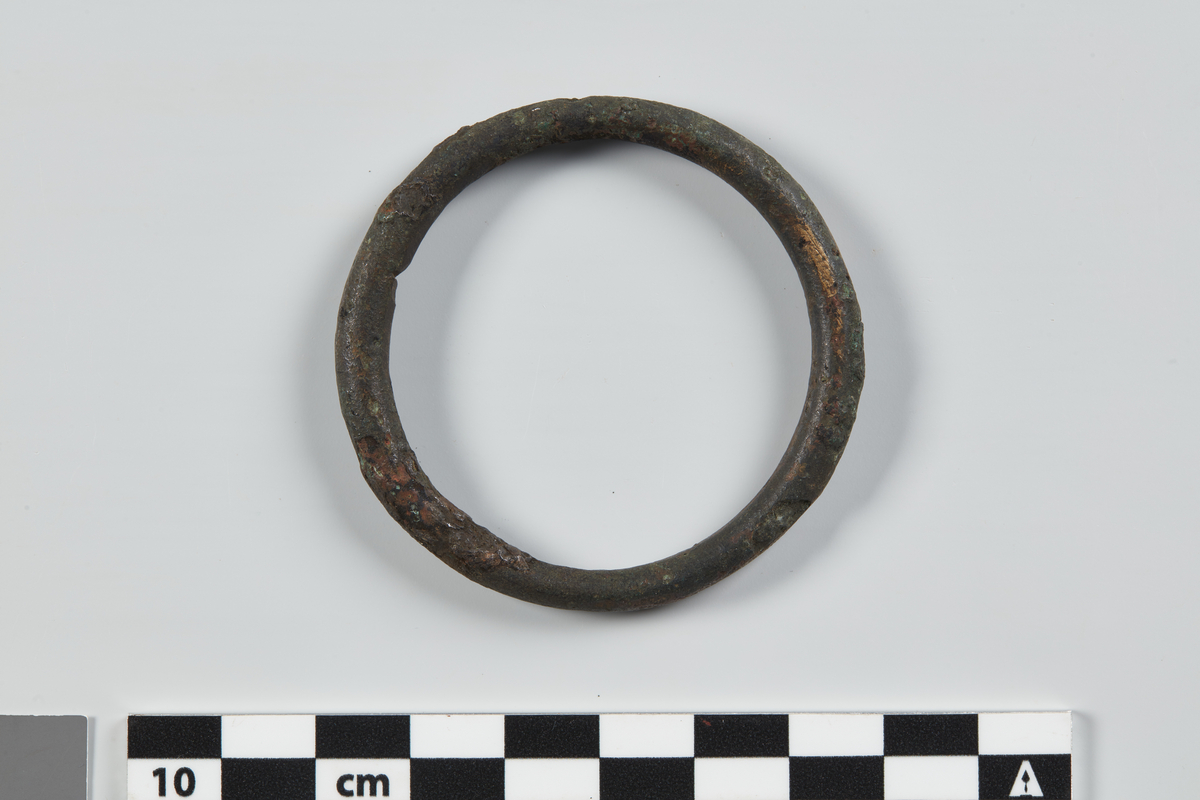 Halvdelen af en flad Ring af Bronce, uden Ornamenter, med en tversover diagnetralt gaaende Broncestang (et Slags Spænde). Ringens tvermaal 2¾"