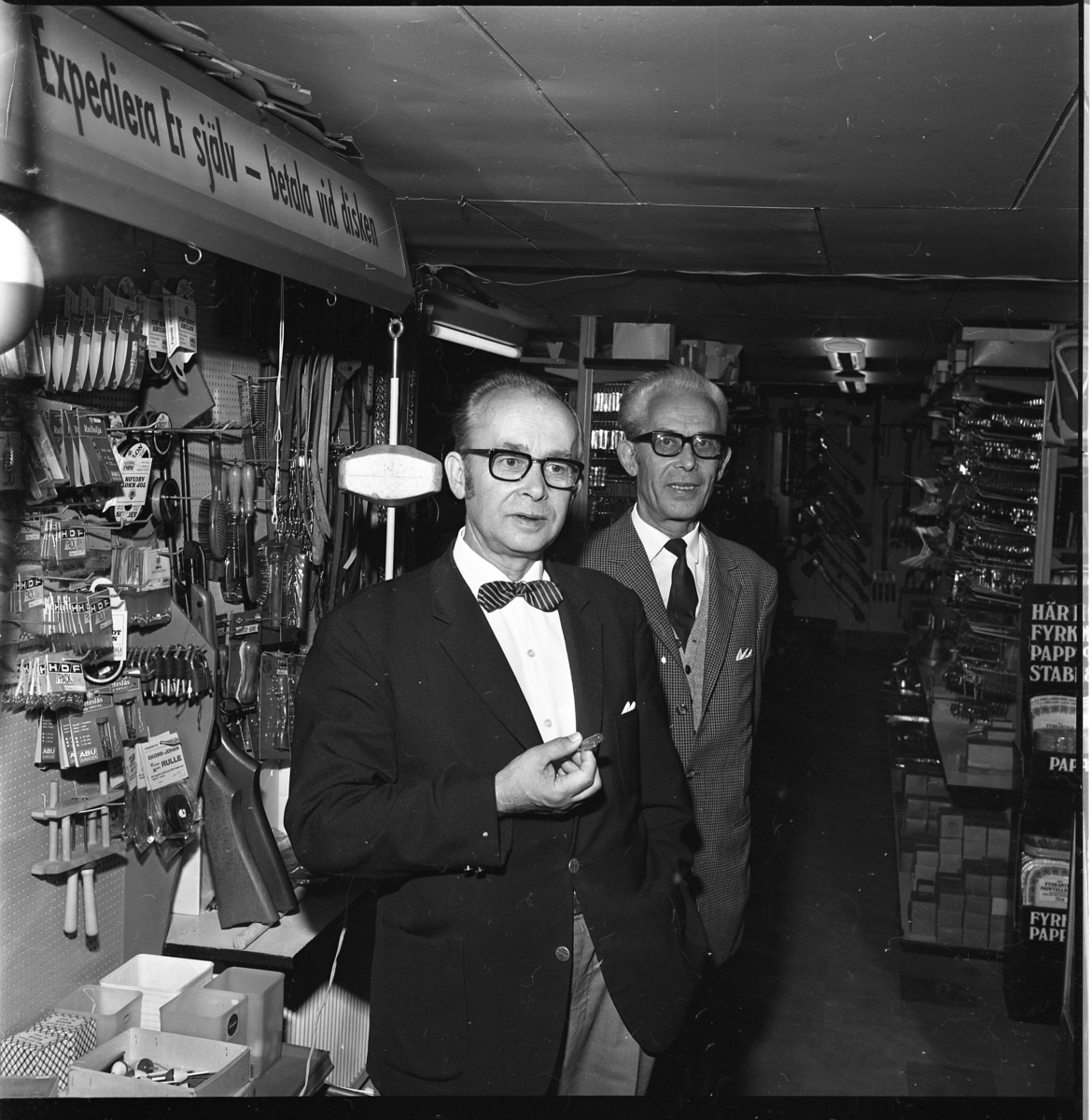 Två välklädda män står i en järnhandel, de bär fluga respektive slips.
