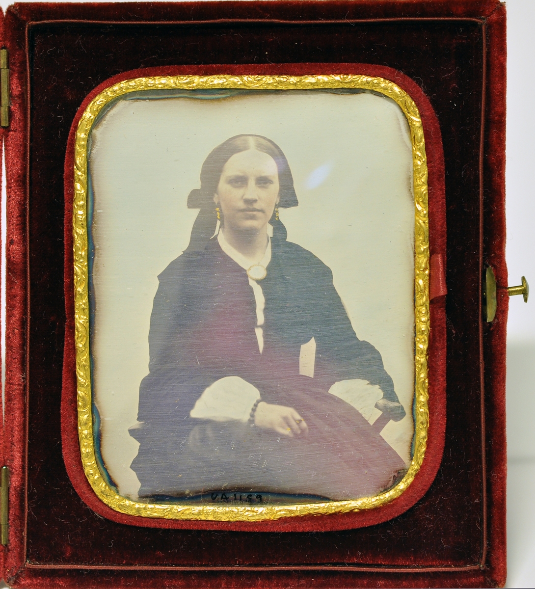 Daguerrotypi i etui. Etui dekket med rød fløyel. To små hengsler og knapp til å åpne. Innvendig portrett av sittende kvinne, gullkant med reileffdekor rundt fotogarfiet, avrundede hjørner. Bildet kan taes ut av etuiet. Kvinnen er Charlotte Aall (1835-1908).