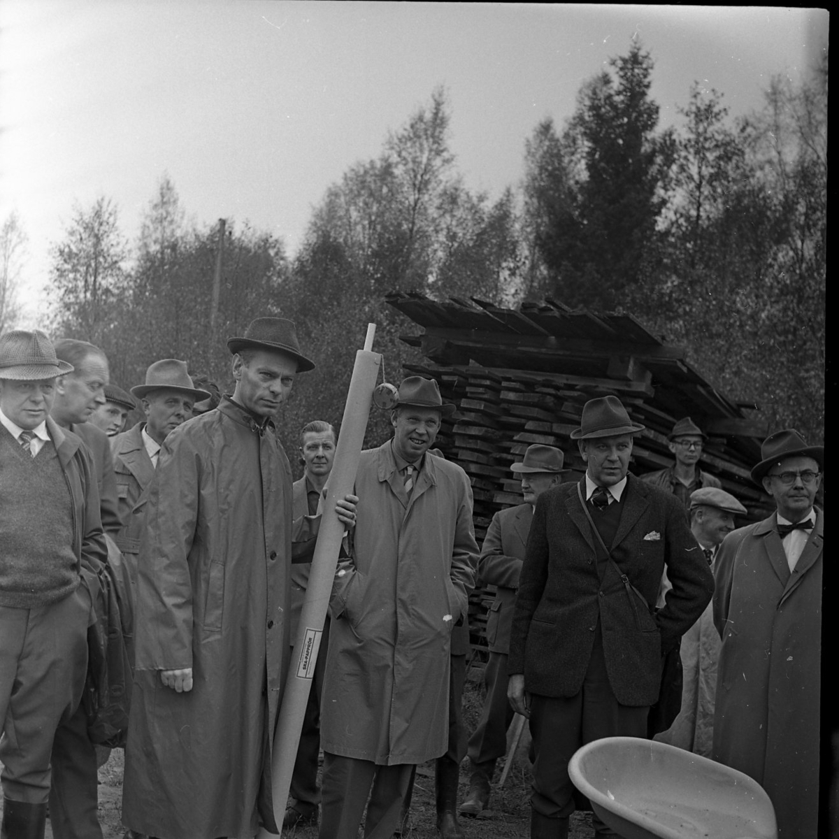 Skogsdag Örserums brunn 1961. Flera män står och tittar på någonting utanför bild. De bär rockar eller regnjackor. Bakom dem står en stapel med bräder.