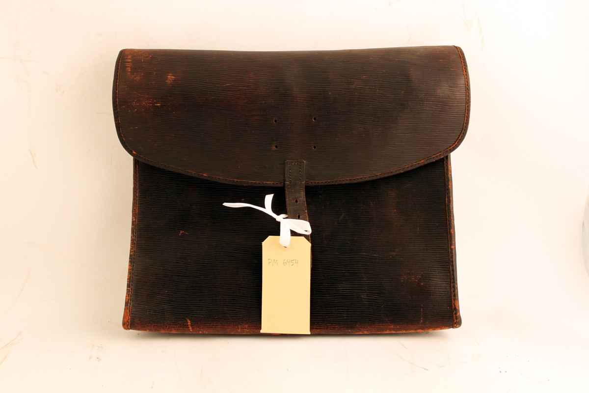 Väska av svärtat läder med axelrem och låsanordning med fastnitade järnöglor och låsten. Väskan försluts med hänglås (saknas). Klaff med förslutning med rem och hålspänne. Fyra hål på klaffen där en bricka varit fästad.