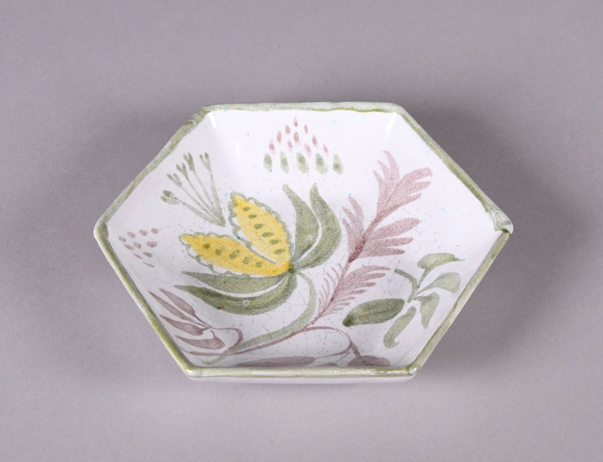 Skål av glassert keramikk, med opphøyd rand. Skålen er sekskantet, og har hvit grunnfarge. Håndmalt dekor av blomster og bladverk, og en grønn borde langs randen.