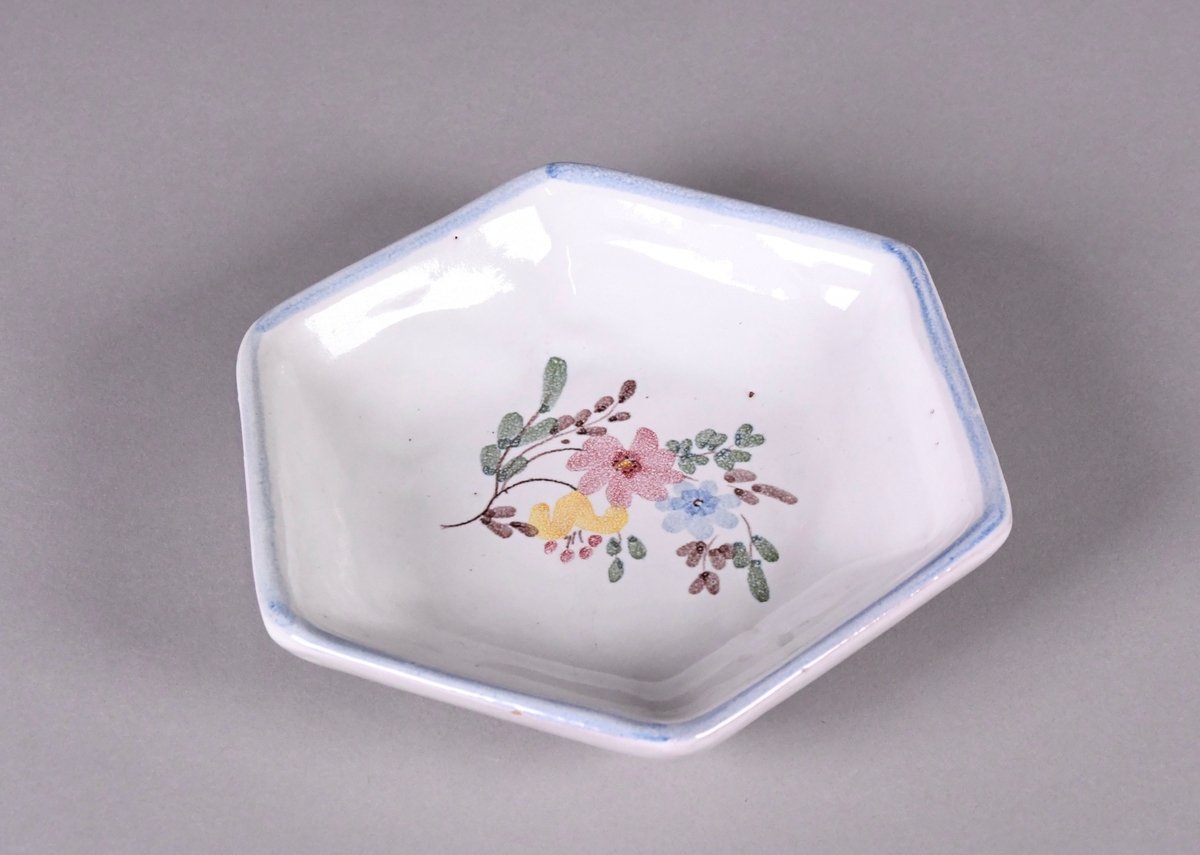Skål av glassert keramikk, med opphøyd rand. Skålen er sekskantet, og har hvit grunnfarge. Håndmalt dekor av gule, røde og blå blomster, og en blå borde langs randen.