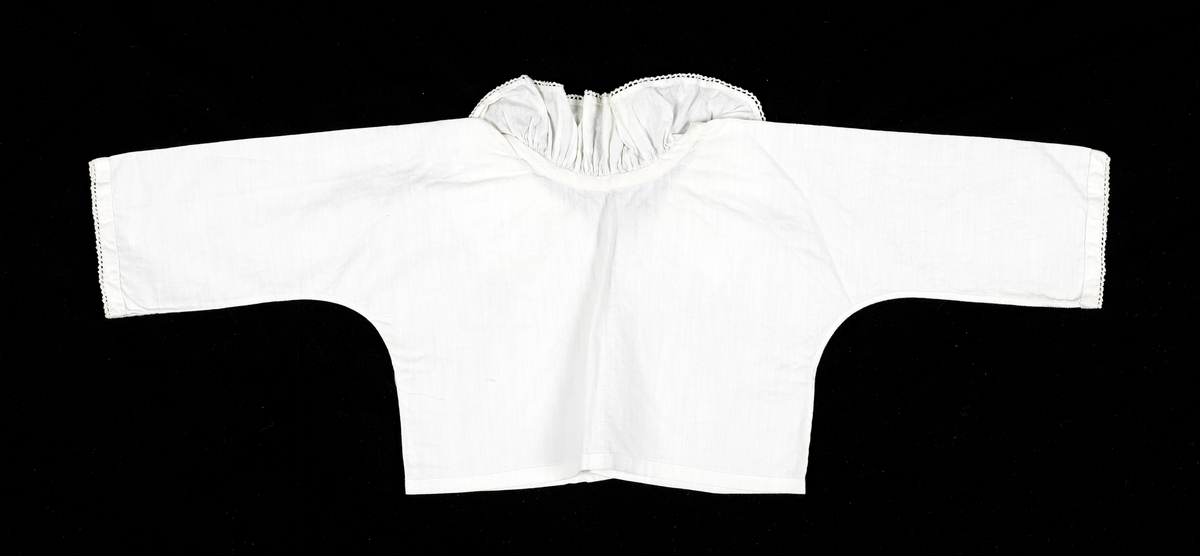 Hvit barneskjorte i bomull med snøring i halsen og bred rynket krage. Heklete detaljer på kant av ermer og krage. Klippet i ett stykke.