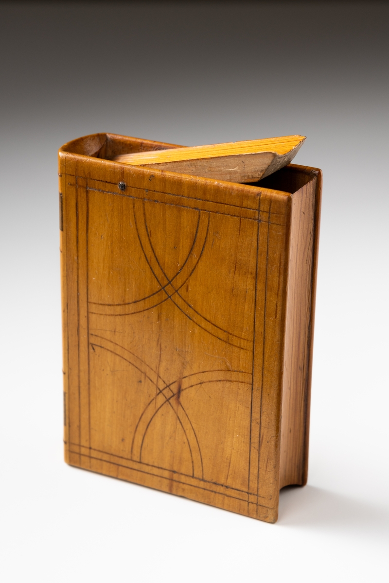 Sparbössa av trä i form av en bok dekorerad med ristat mönster och intarsia. På ovansidan av bössan finns en fällbar lucka innanför vilken myntinkastet är placerat. Skramlande föremål invändigt.