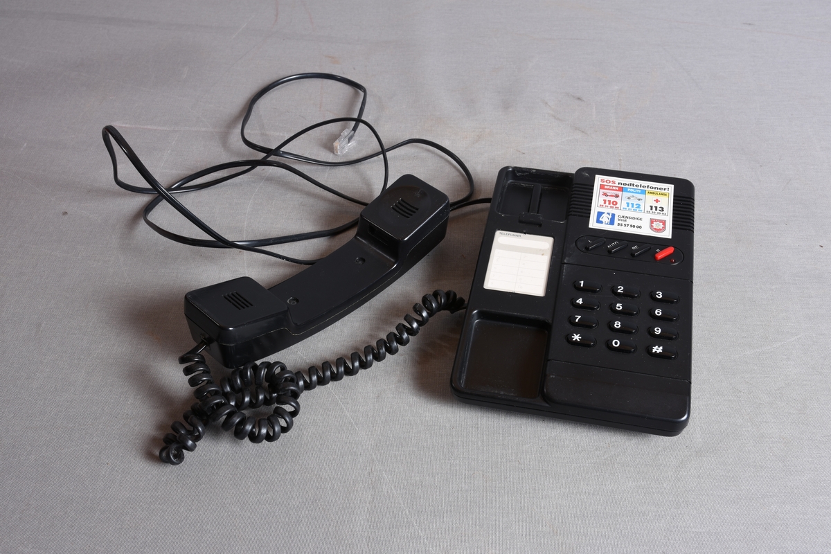 Svart fasttelefon med base, rør med ledning, samt tilkoblingskontakt.