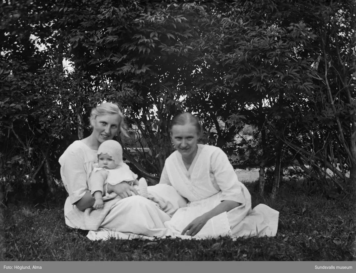 Småskollärarinnan Anna Höglund med en baby i famnen samt en annan kvinna sittandes i gröngräset.