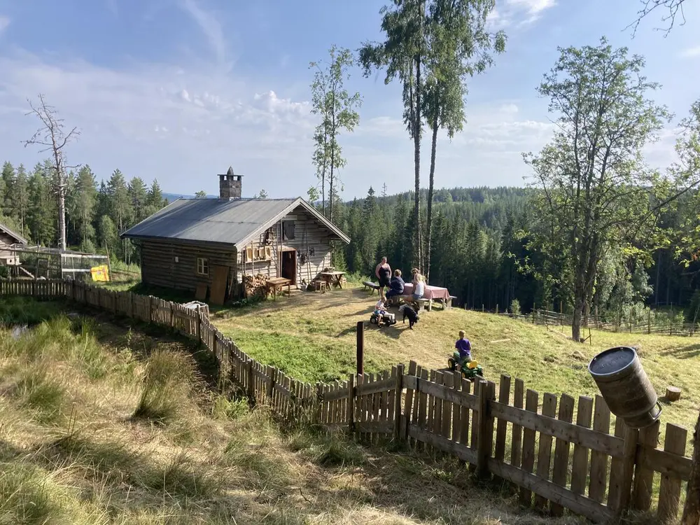 Oversiktsbilde over Skålbergsætra i fint sommervær. Sætra ligger bak et gjerde. Foran sætra sitter noen mennesker ved et bord og koser seg i sola.