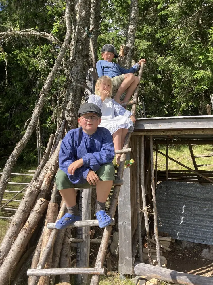 Tre barn sitter på en trestige lent mot et tre og et uthus.