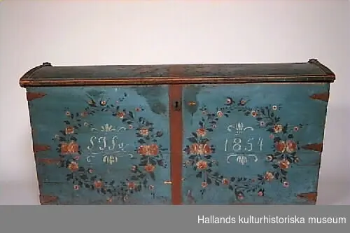 Blåmålad kista av trä med två blomsterkransar som dekorerar kistans framsida. Blommorna i kransarna utgörs av vit-rosa rosor samt mindre enkla blommor i liknande färg och gröna blad. Även locket dekorerat med en båge av liknande blommor. I ena kransen monogrammet "SJS", i andra kransen årtalet "1854".