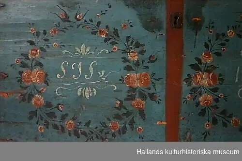 Blåmålad kista av trä med två blomsterkransar som dekorerar kistans framsida. Blommorna i kransarna utgörs av vit-rosa rosor samt mindre enkla blommor i liknande färg och gröna blad. Även locket dekorerat med en båge av liknande blommor. I ena kransen monogrammet "SJS", i andra kransen årtalet "1854".