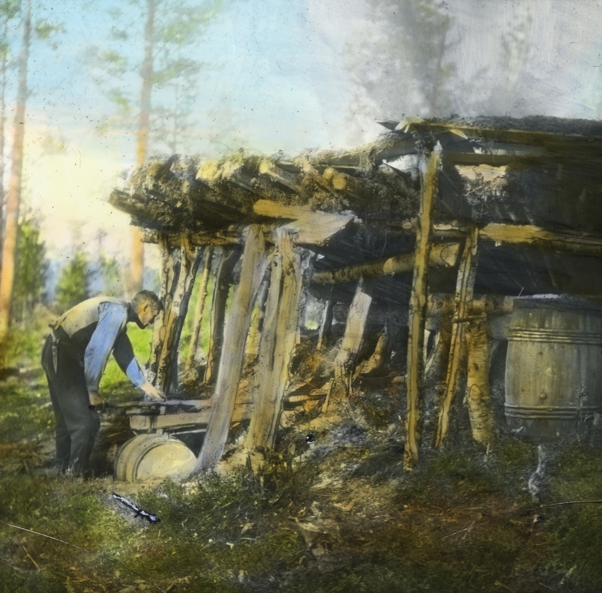 Håndkolorert dias. Tjærebrenning i Målselvdalen i 1928. En mann kontrollerer tappingen av tjære på tønner fra mila.