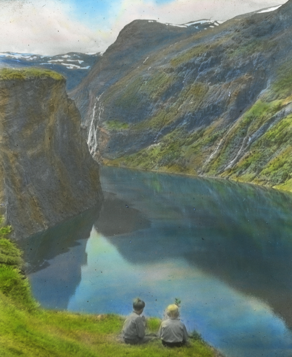 Håndkolorert dias. To barn sitter på en gresskledd bakke og skuer utover Geirangerfjorden.