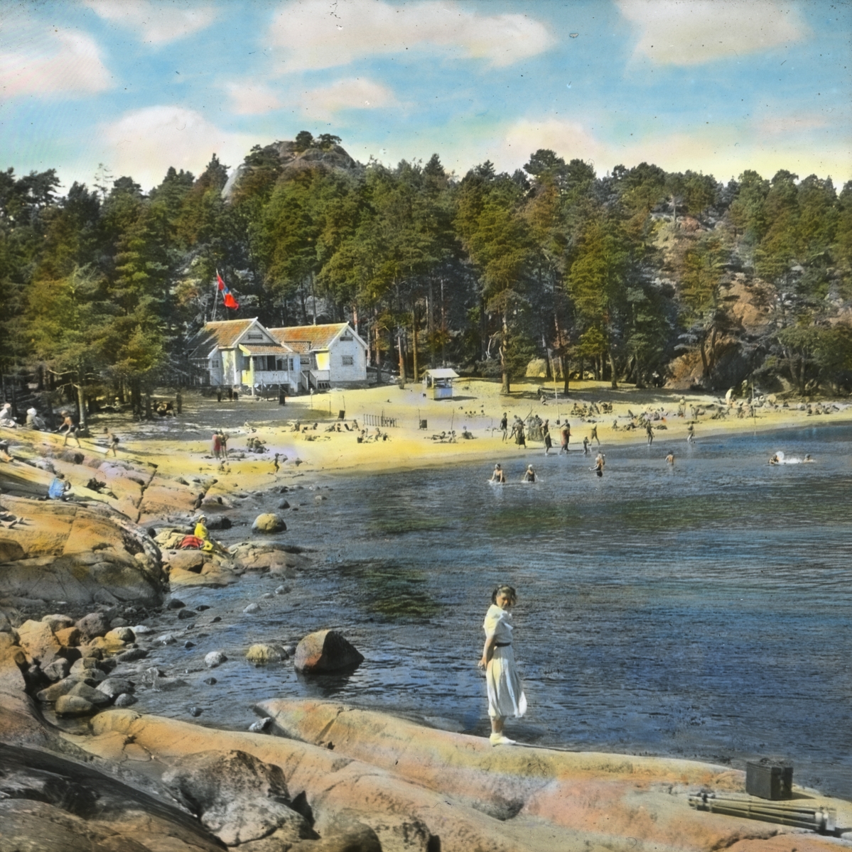 Håndkolorert dias. På bildet ser vi badende mennesker på Storesand strand i Grimstad. I bakgrunnen ser man skog og to hvite hus. En ung kvinne står på svaberget i forgrunnen.