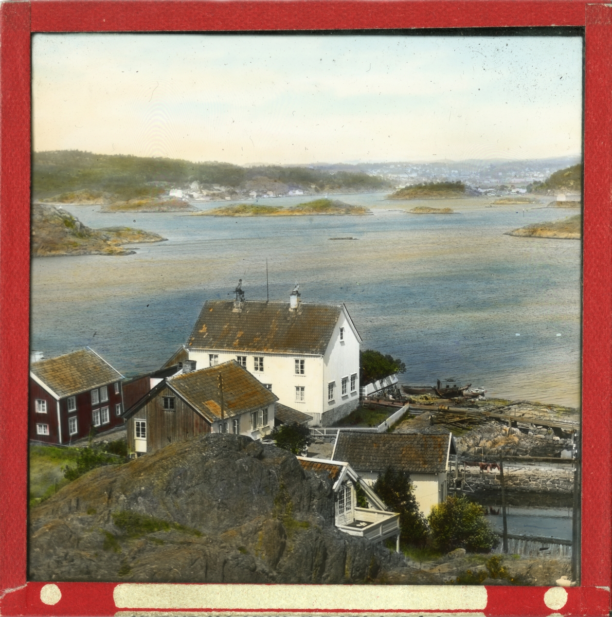 Håndkolorert dias. Utsikt fra Merdø mot Arendal. En husklynge med våningshus, bryggerhus og båthus ligger ved vannkanten.