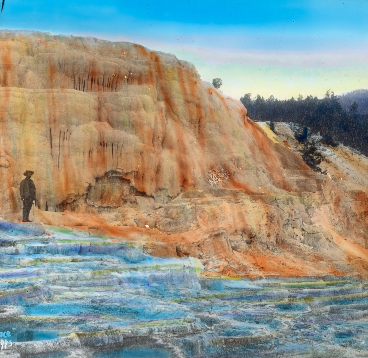 Håndkolorert dias. En mann står i Mammoth Hot Springs, som er et stort område med varme kilder i nasjonalparken Yellowstone i U.S.A. Formasjonene har vokst frem over tusenvis av år ved at varmt vann fra kildene er blitt avkjølt og har dannet kalkstein.