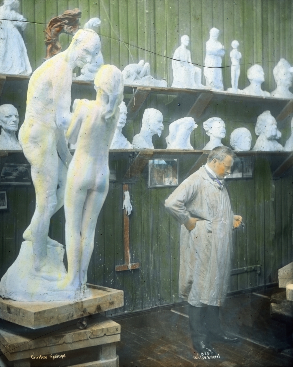 Håndkolorert dias. Portrett av billedhuggeren Gustav Vigeland i hans atelier. Bak Vigeland står en rekke skulpturmodeller og byster. I forgrunnen står en skulpturgruppe av en eldre mann og en yngre jente.
