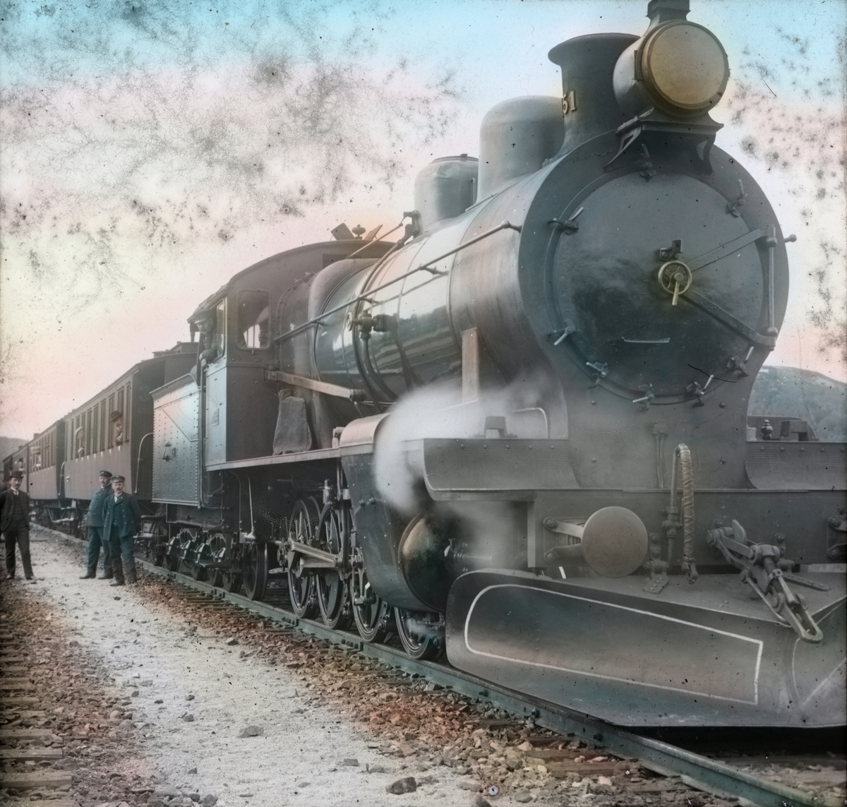 Håndkolorert dias. Et damplokomotiv med en lang rekke vogner. I bakgrunnen sees tre menn ved siden av toget.