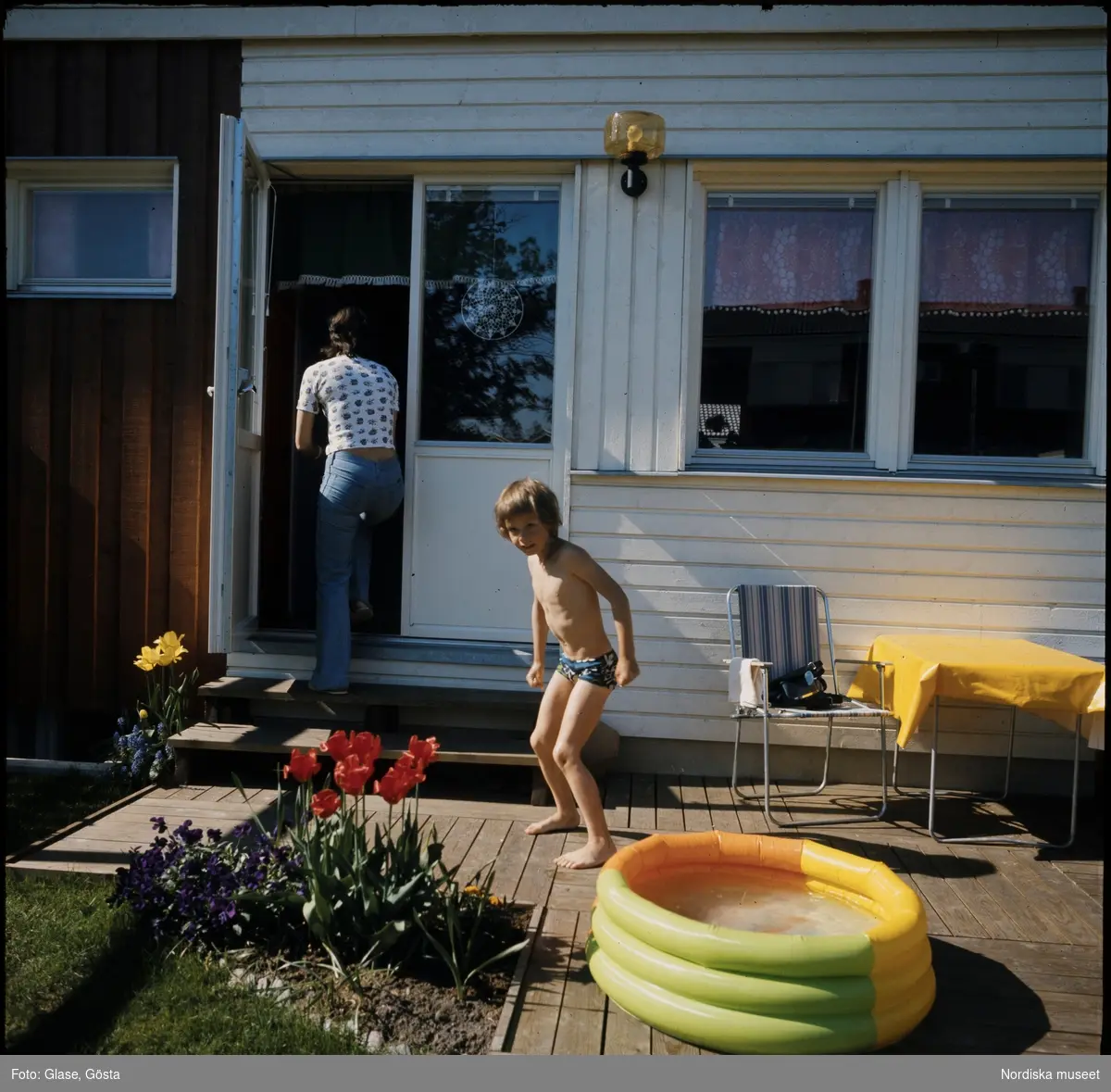 Uppblåsbar pool på radhusaltan. Ett barn står berett att bada, en kvinna påväg in i huset.