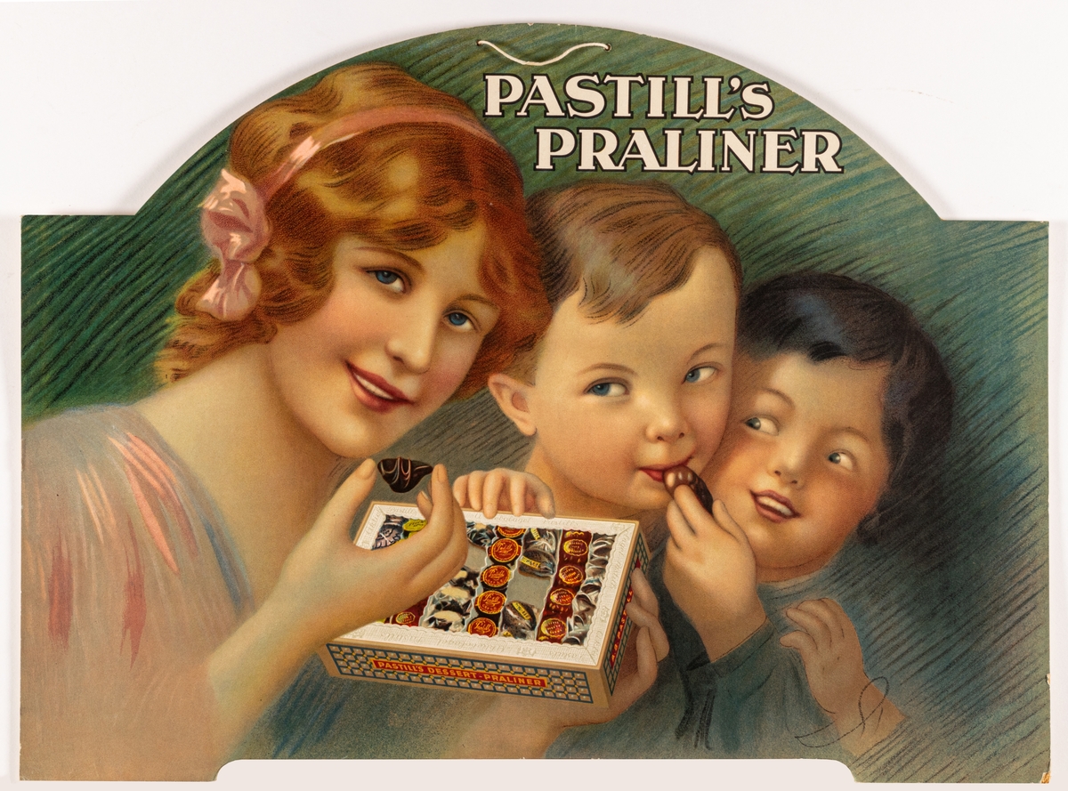Reklamskylt av papp, flerfärgstryck, för Pastill's praliner. Uppställningsbar med flik på baksidan och upphängningssnöre. Kvinna och två barn håller en pralinask, grön bakgrund.