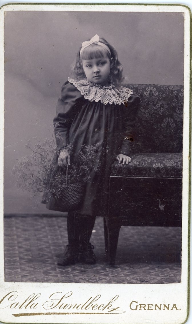 Kabinettsfotografi av en okänd liten flicka med rosett i håret och en korg med blommor i handen. Hon står intill en soffa.