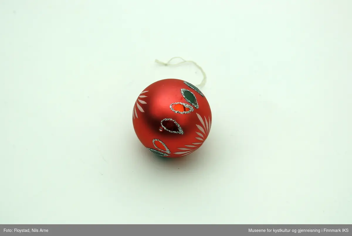 En liten julekule i hard plast med en halvmatt rød/mørkrosa farge med dekor. Dekoren består av påmalte hvite striper på begge sider av kulen og har et blomsterlignende mønster. I tillegg er det dråpeformet dekor malt rød og grønn med glitterkant som går vertikalt rundt kulen, og danner tilsammen en linje. Julekulen har et oppheng av metall med en hvit bomullstråd. Opphenget har gravert mønsterdekor.