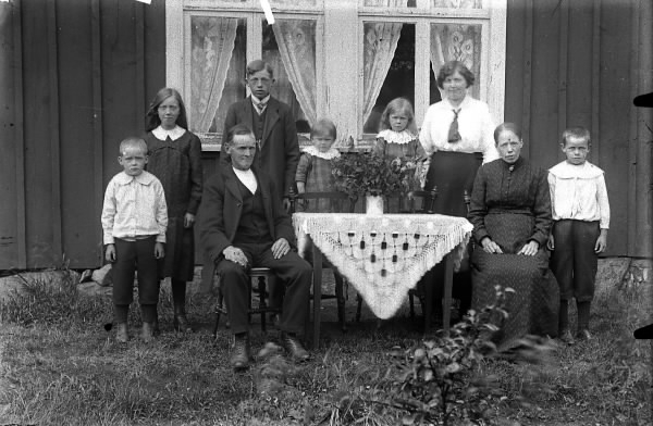 Familjebild vid ett bord utanför ett par av bostadhusets fönster. Tre vuxna och sex barn.