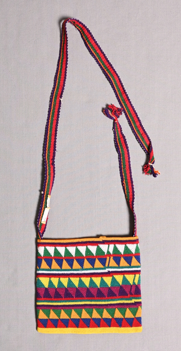 Mönstervirkad väska från Nebaj, Guatemala. Vävt band/bärrem i randig varprips. Bärremmen i 2 delar med tofs i var ände. Väskan med geometriskt mönster i vitt, blått, orange, grönt, rött, vinrött, lila, och gult.