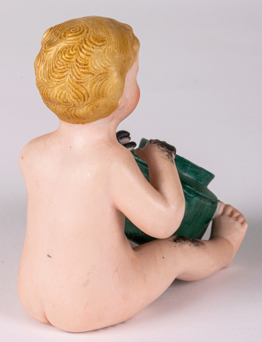 Bläckhorn i form av en naken pojke som sitten med en grön burk framför sig. Pojken är blond med svarata händer och bläckfläckar i ansiktet. Ihålig, av keramik.