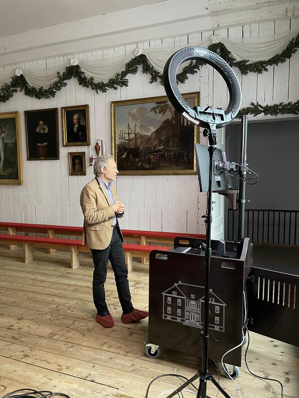 Torleif Hamre fra Eidsvoll 1814 har et digitalt møte med en klasse i Sandefjord. Torleif står i Rikssalen ved en  vogn på montert kamera, skjermer, og lys - et mobilt enmanns TV-studio.