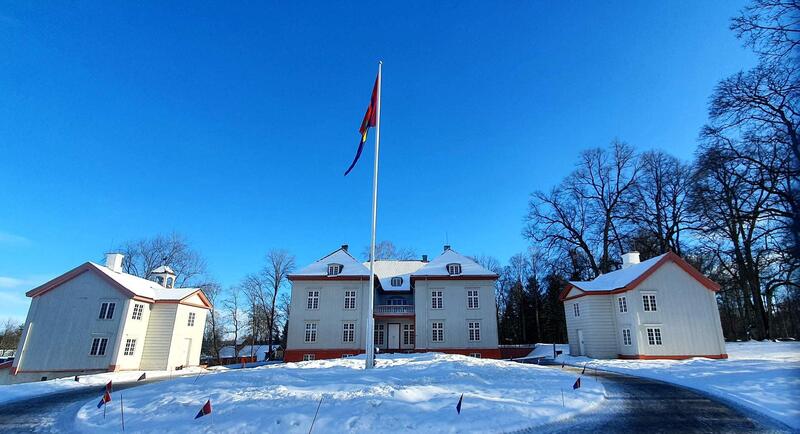 Et stort oversiktsbilde av Eidsvollsbygningen og sidebygningene en vakker vinterdag. Snø på bakken, knallblå himmel og samisk flagg i flaggstanga foran bygnignen