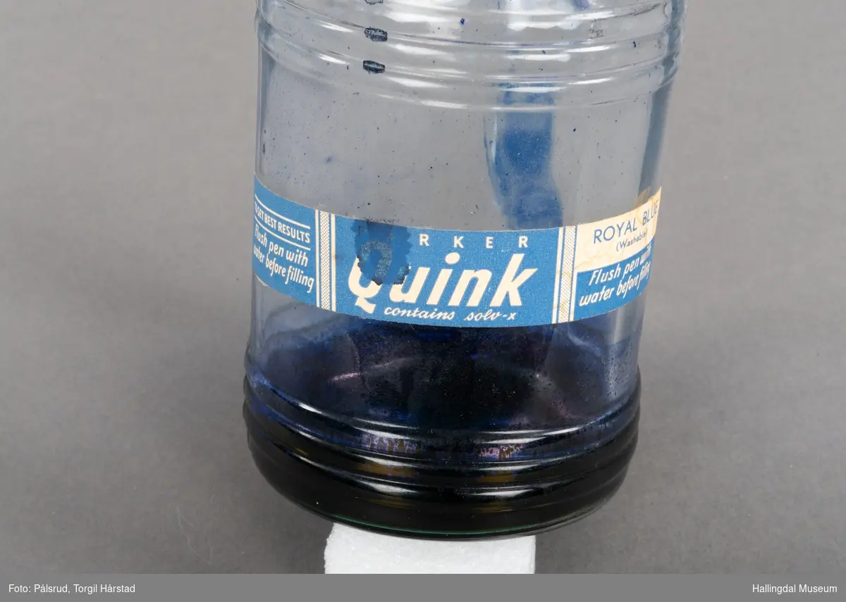 Flaske av blankt glas. Rett, sirkulært tverrsnitt. Det er litt blekk på flaska. Skruvekork av svart plast.
