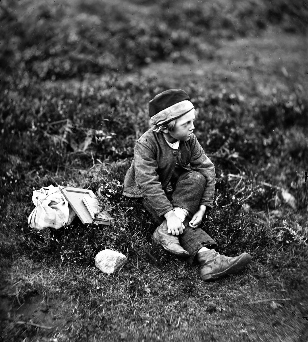 Pojke sitter i gräset med skolböcker och griffeltavla. Severins systerson Karl Nilson.