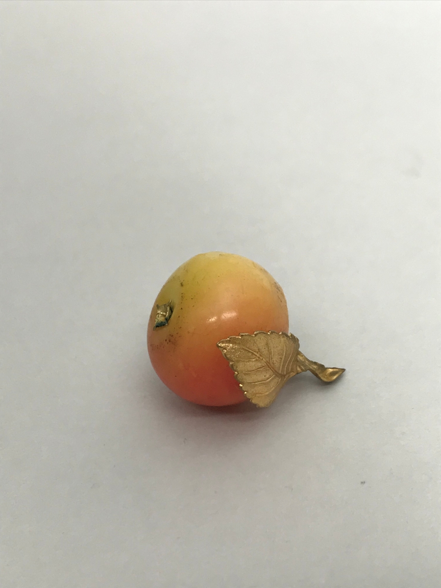 Lite eple av voks, rølig på den ene siden, gulere på den andre. Blad og stilk av forgylt metall. Eplet har sannsynlig vis vært del av den større dekorgjenstand?