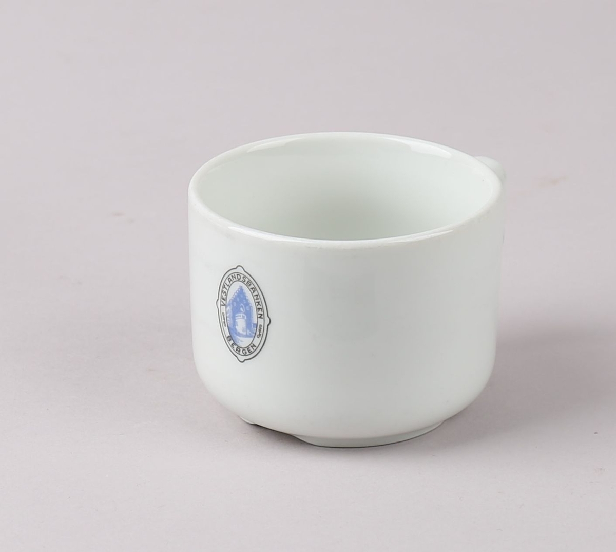Kaffekopp, del av kaffeservise, 1 kuvert. Koppen har sylindrisk form. Produsert i hvit porselen med emblem på korpus, for Vestlandsbanken i Bergen, med tegning av Håkonshallen i emblemet.
