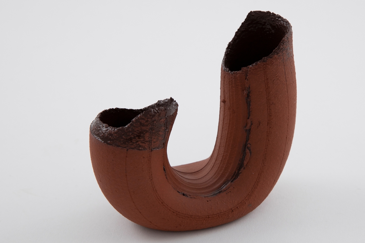 Vase i matt brun terrakotta. Asymmetrisk U-form med riller. Åpne munninger med ujevne render. Mørkebrunglasert på innsiden.