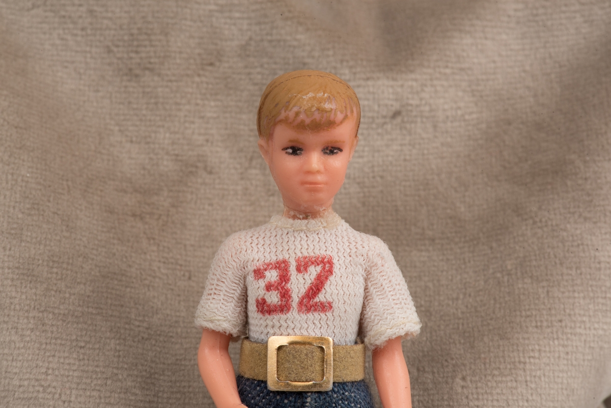 Pojkdocka för dockskåp i plast, troligen tillverkad av Lundby leksaksfabrik.
Dockan är klädd i kortärmad vit tröja med ett tryck i rött på framsidan "32".  Blåa långbyxor med brunt skärp och guldspänne.
På fötterna ditmålade svarta skor.