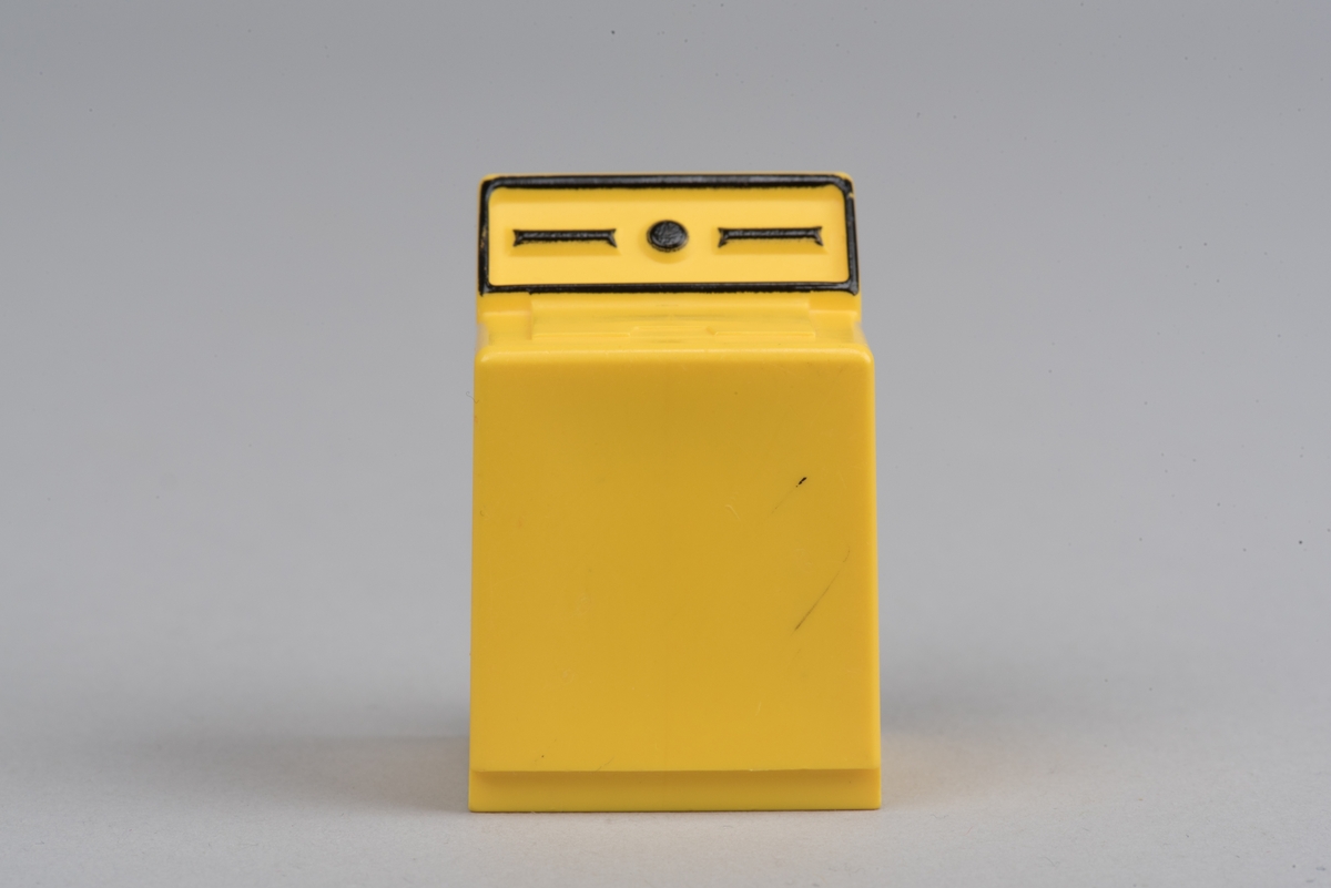 Dockskåpsinredning i form av en torktumlare av  gul plast.
Torktumlaren har en lucka på ovansidan i svag relief. På det förhöjda bakstycket finns en "knapp" och två längsgående ränder inom en ram, allt i svart och i relief.