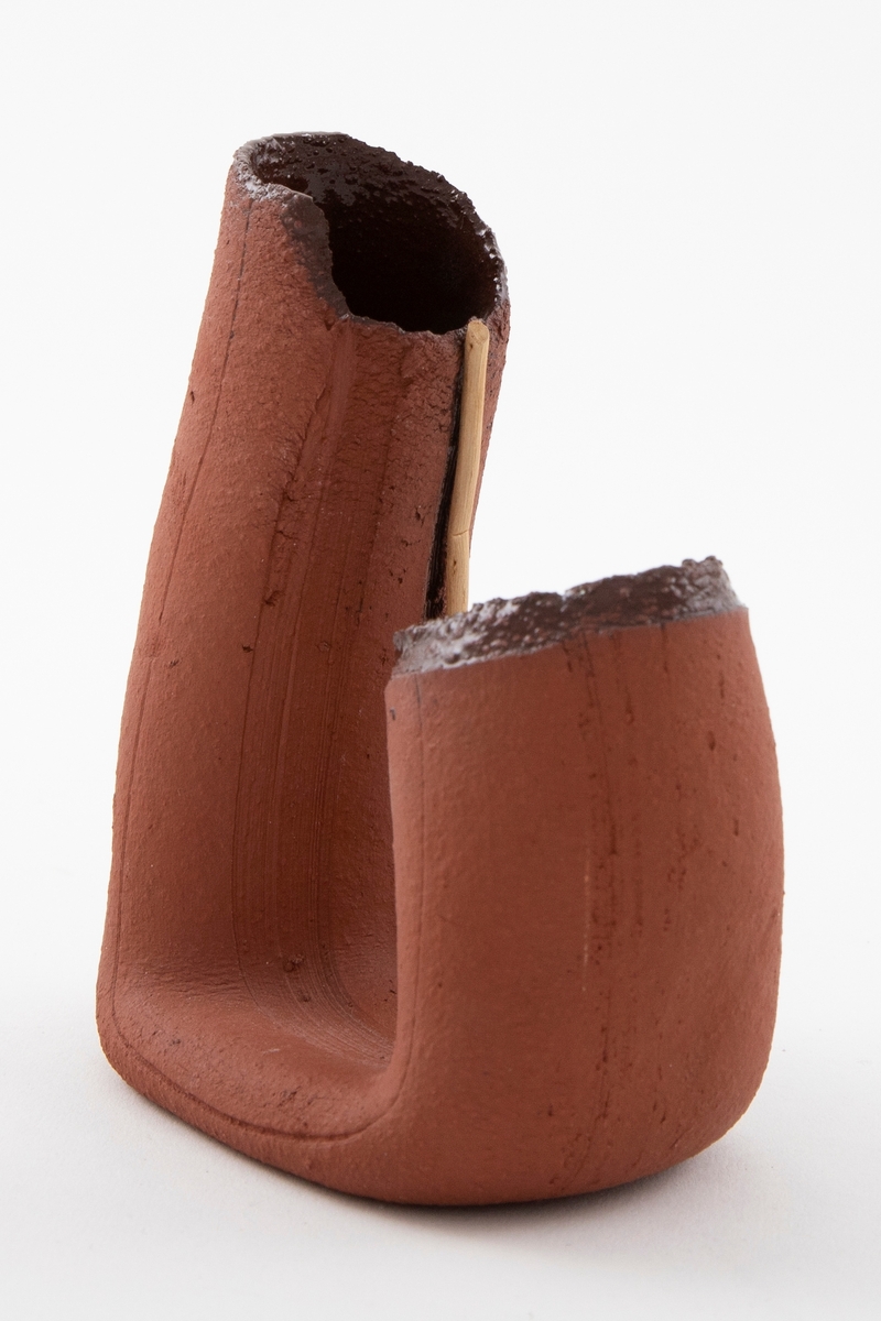 Matt brun vase i terrakotta. Asymmetrisk U-form med åpne ender med ruglete render. En bjørkerot ligger på innsiden av U-formen og følger den. Vasen er blank brunglasert på innsiden.