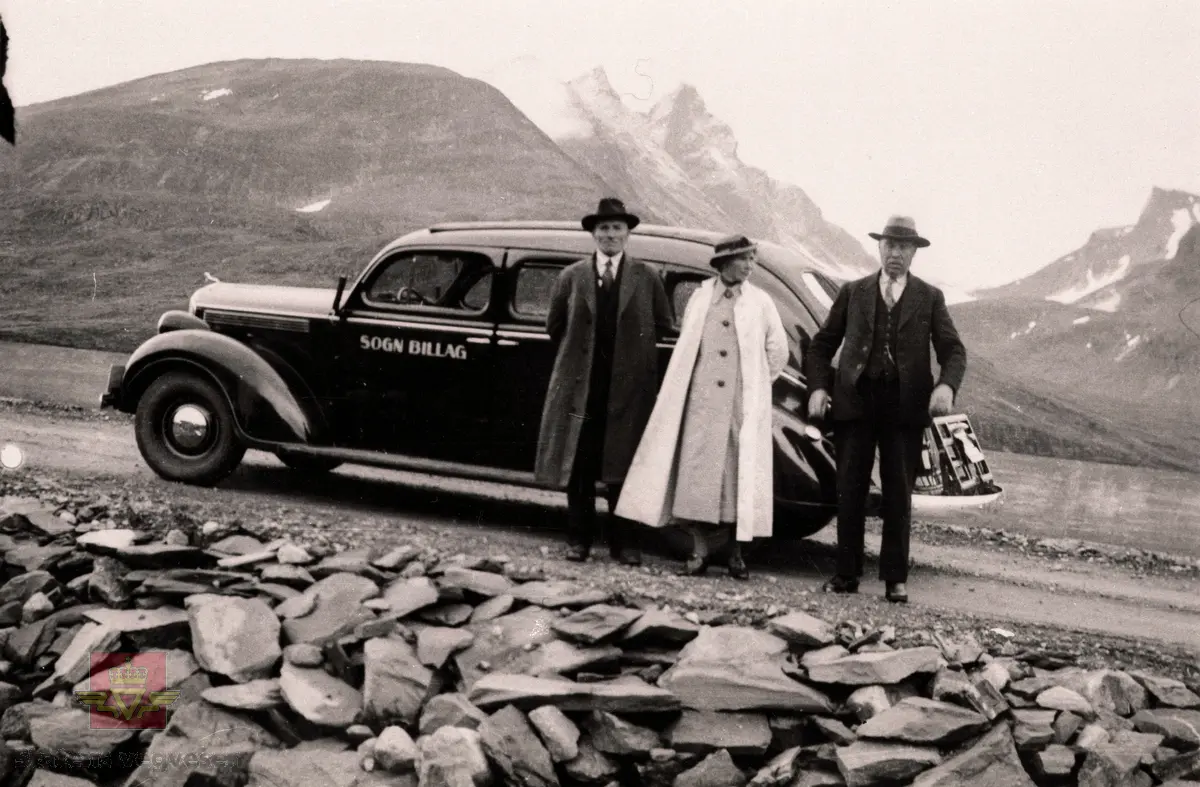 1938 modell Dodge rutevogn på riksveg 55 Sognefjellet.  

Rutevogna er merka "Sogn Billag". Frå venstre: Hans Grinde, styremedlem i Sogn Billagg L/L , til høgre er banksjef Tefre og mellom står fru Tefre.