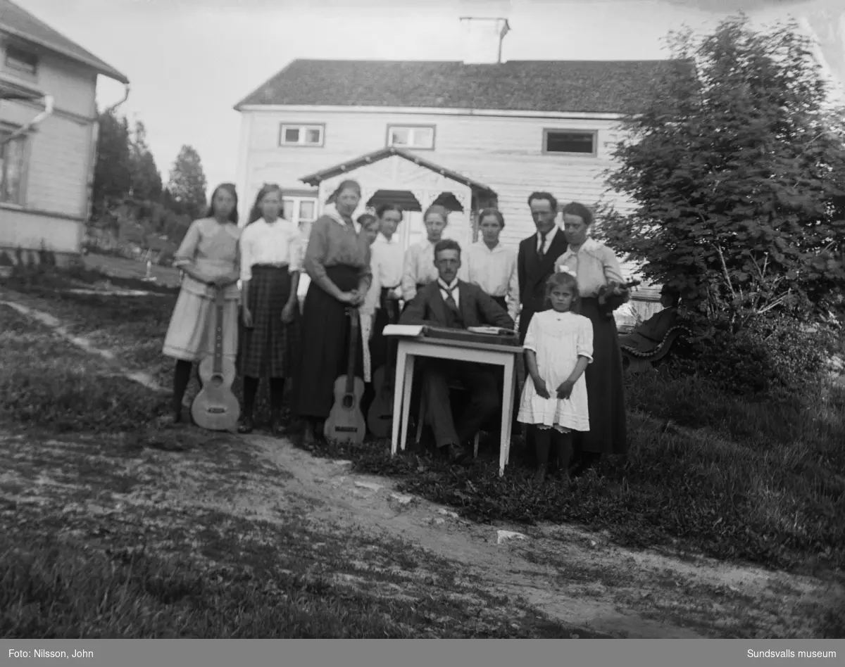 Två gruppfoton från gårdstunet på Roken 1:7 i Laggarberg. Vuxna och barn, många av dem med musikinstrument, en man sitter vid ett litet bord i mitten. På första bilden syns även en man som läser tidningen i bakgrunden.