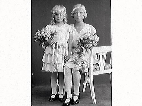 Syskonbild av två flickor i sommarskrud med blomdiadem, volangkjolar och blombuketter. Kamrer Bramstång beställde bilderna, sannolikt barnens far.