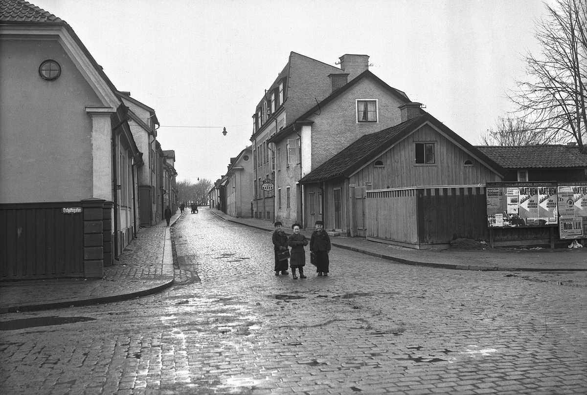 Några barn och fotografen möter ömsesidiga blickar. Platsen är Hunnebergsgatan i Linköping med vy västerut från Östgötagatan. Affischeringen till höger daterar bilden till år 1938.