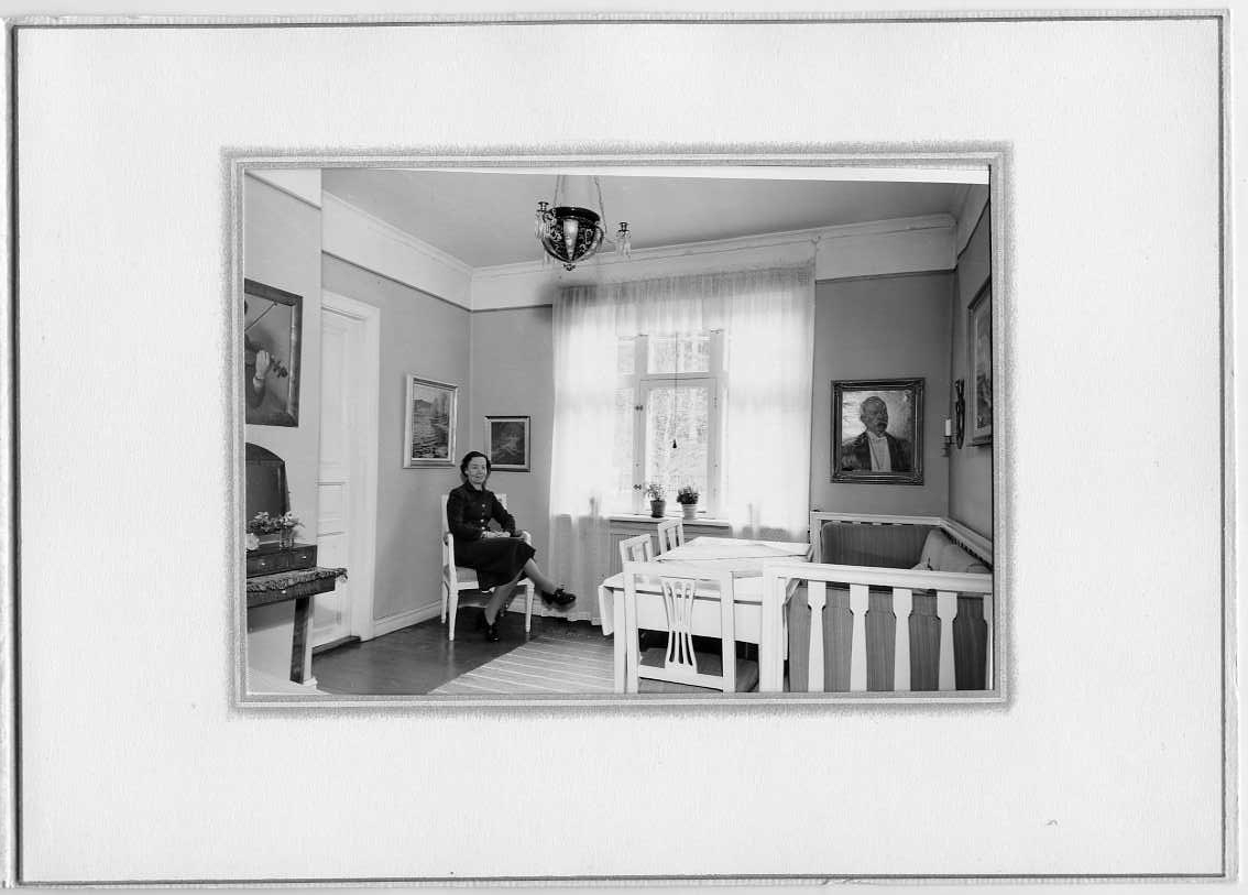 Interiör från fröken Astrid Malmborgs föräldrars hem. Hon sitter på en stol i ett hörn intill ett stort fönster med ljusa, tunna gardiner. Till vänster om henne en stängd dörr och till vänster om densamma hänger det en målning med en fiolspelande man, möjligen målning självporträtt av Edvard Berggren. Mittemot står det en vit soffgrupp med slagbord, stolar och soffa. På väggen hänger flera målningar, bland annat ett porträtt föreställande hennes morfar, borgmästare Victor Egnell i ämbetsuniform. Det är målat av hennes pappa, konstnären Pelle Malmborg.