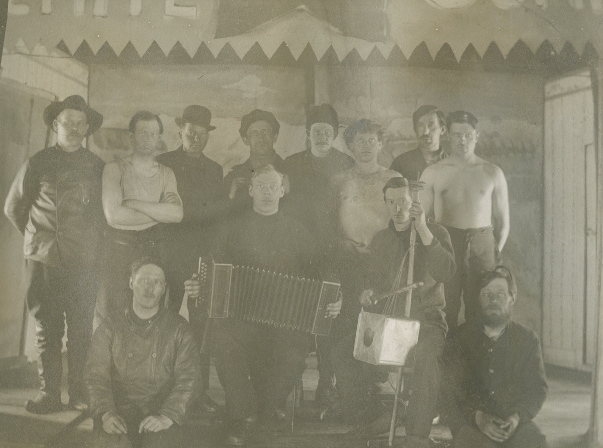 Fotografi från expedition till Spetsbergen. Motiv av en grupp män som poserar på vad som liknar en scen. Några av dem bär hatt, andra är barbröstade. Två av dem håller i instrument, ett dragspel och ett stränginstrument.