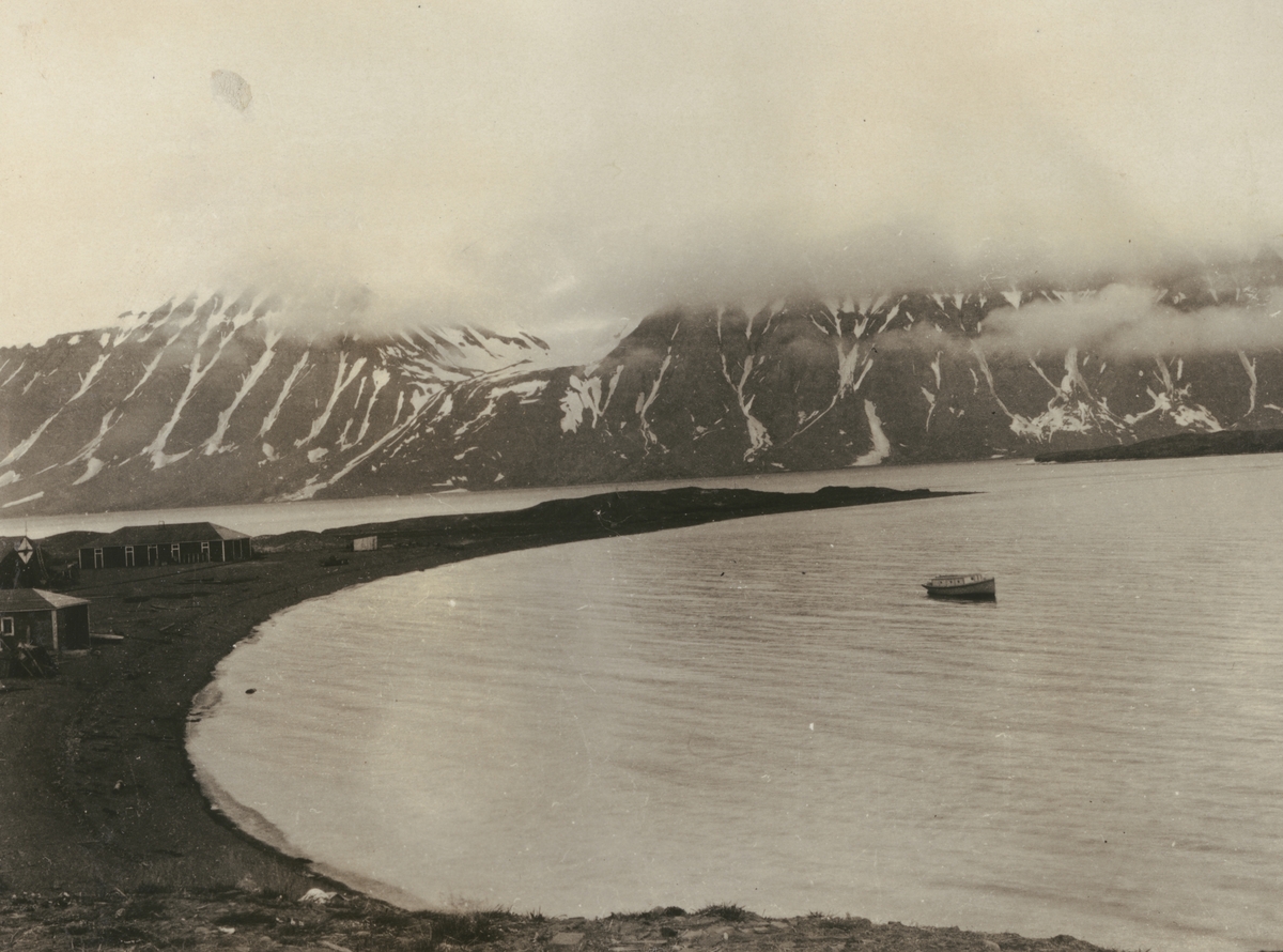Fotografi från expedition till Sveagruvan. Vy över havsvik med snötäckta berg i bakgrunden. På stranden syns några hus.